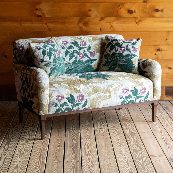 Angled front view of a settee upholstered in a floral fabric with two matching throw pillows against a wooden wall 