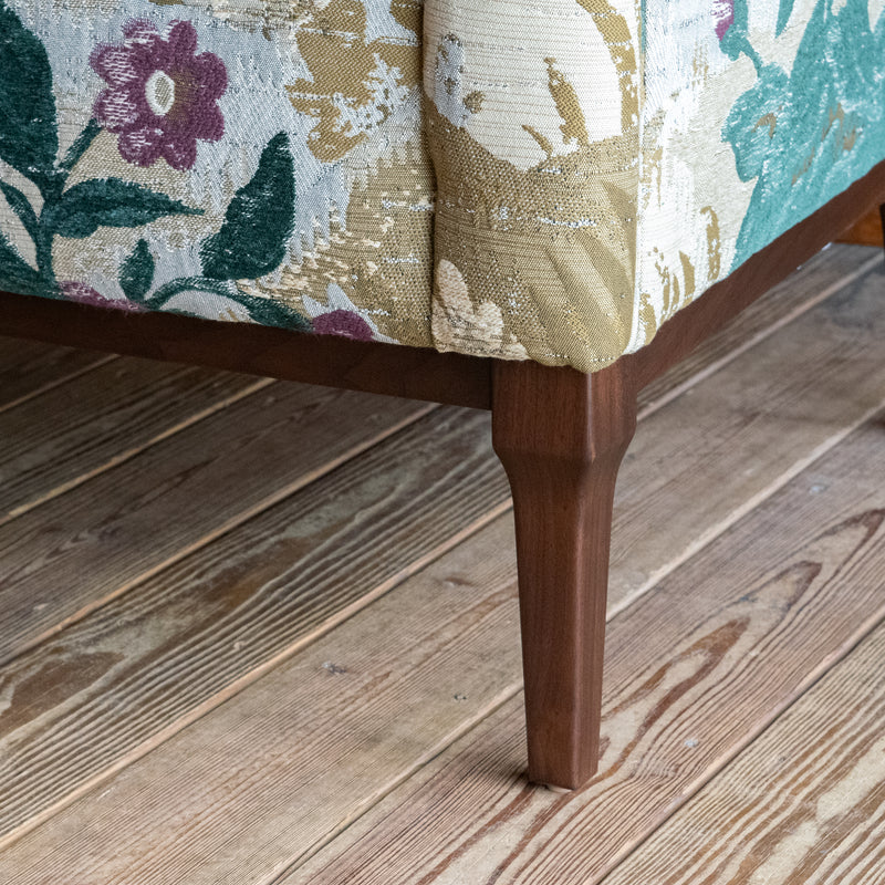 Close-up of the leg of a settee upholstered in a floral fabric against a wooden floor