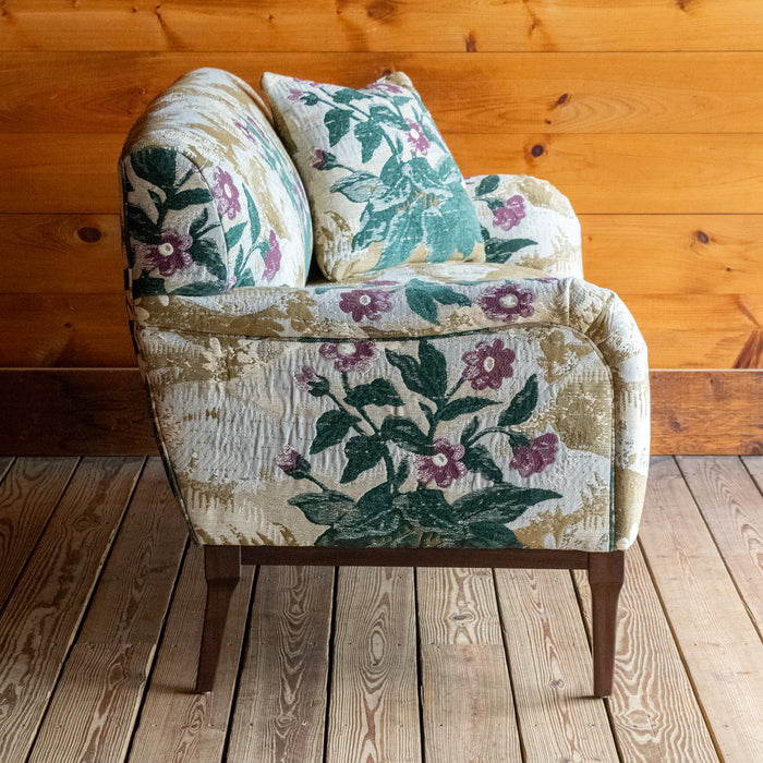 Profile view of a settee upholstered in a floral fabric with two matching throw pillows against a wooden wall 