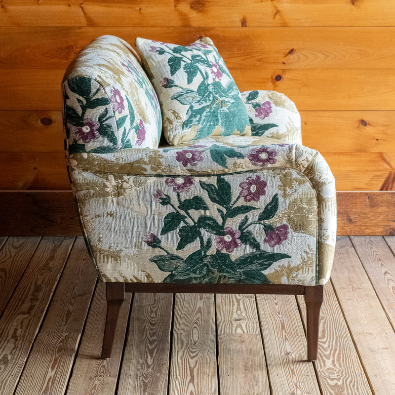Profile view of a settee upholstered in a floral fabric with two matching throw pillows against a wooden wall 