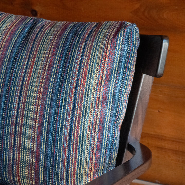 Angled back view of an exposed wood frame chair with striped cushions against a wooden wall