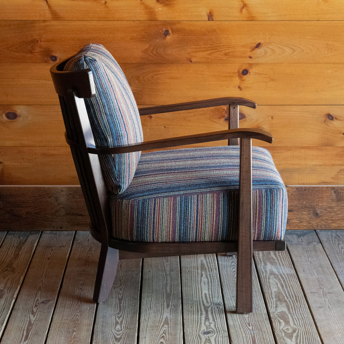 Profile view of an exposed wood frame chair with striped cushions against a wooden wall
