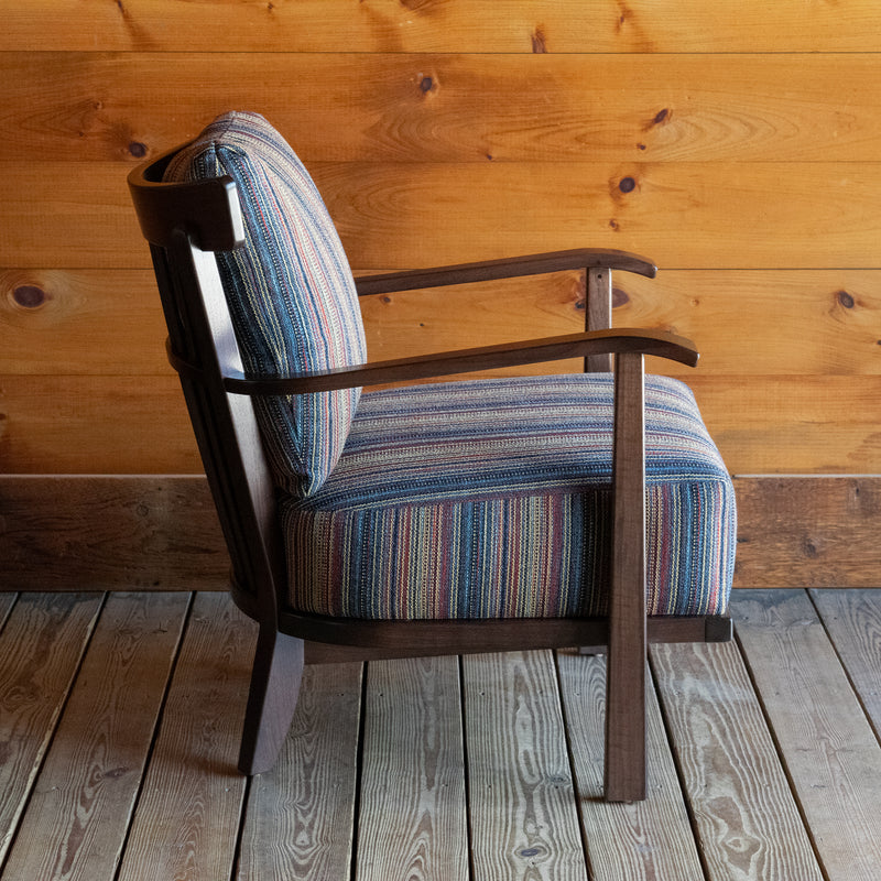 Profile view of an exposed wood frame chair with striped cushions against a wooden wall