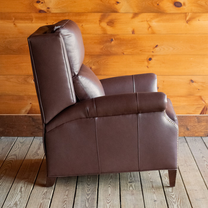 Profile view of a dark brown leather recliner with rolled arms and nailhead trim against a wooden wall