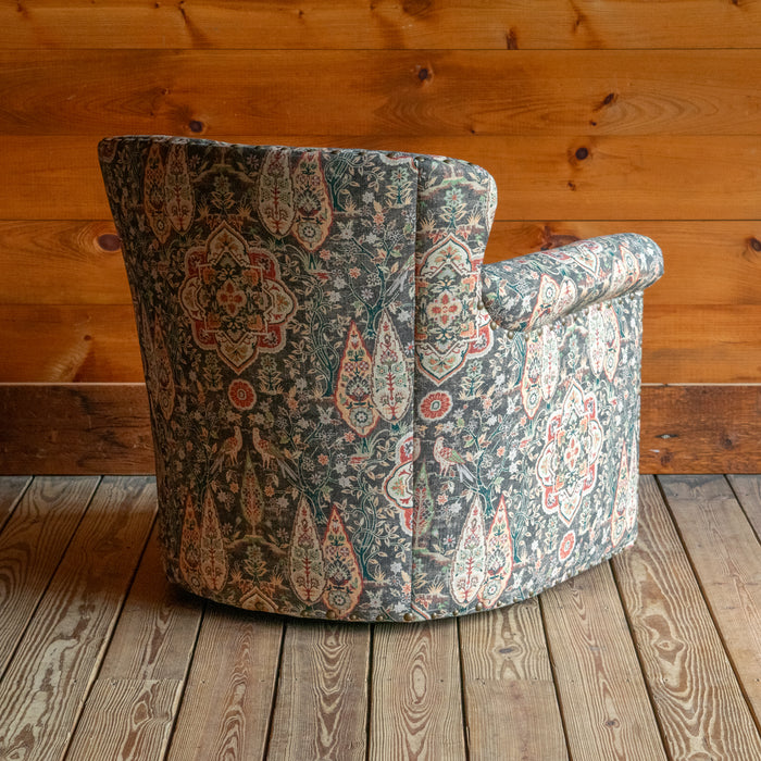 Angled back view of a swivel chair upholstered in muted sage green fabric with bird and floral motifs against a wooden wall 