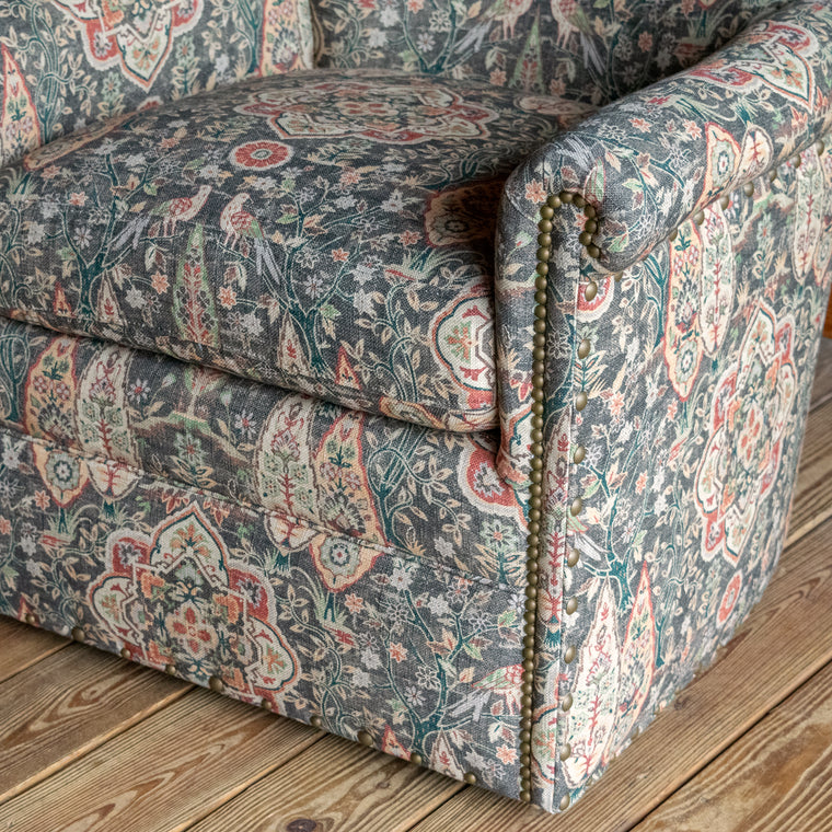 Angled back view of a swivel chair upholstered in muted sage green fabric with bird and floral motifs against a wooden wall 