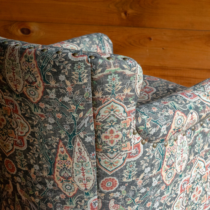 Back corner close-up of a swivel chair upholstered in muted sage green fabric with bird and floral motifs against a wooden wall 