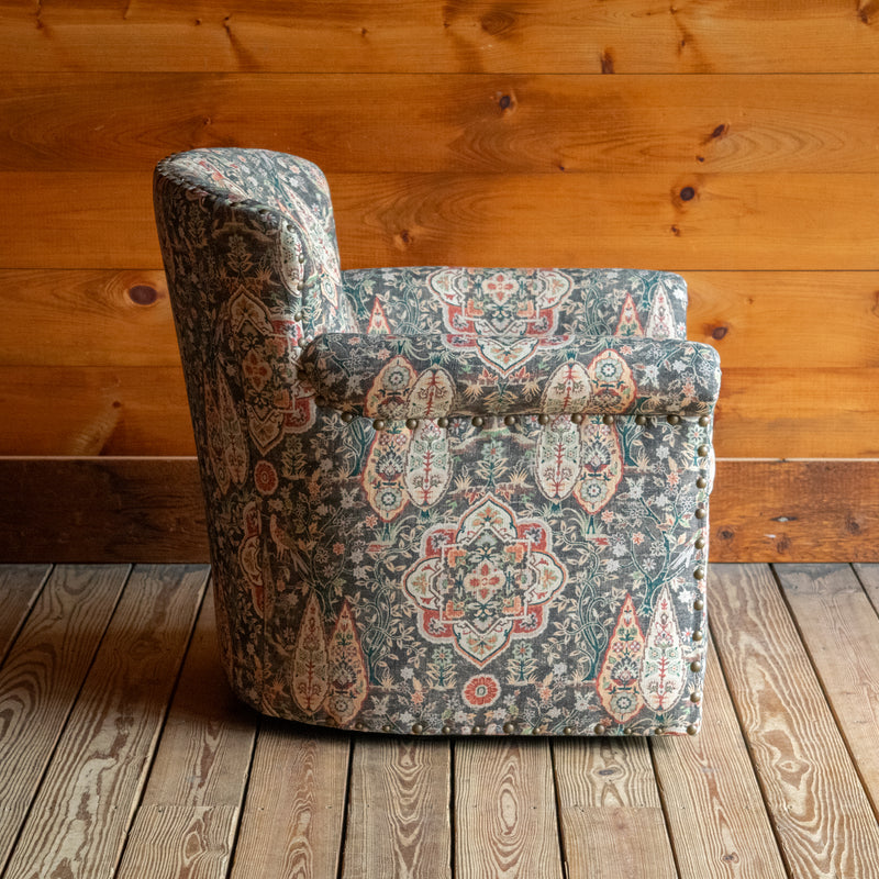 Profile view of a swivel chair upholstered in muted sage green fabric with bird and floral motifs against a wooden wall 