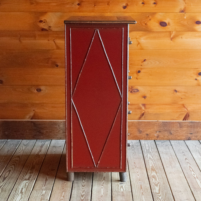 Profile view of tall, red painted four-drawer dresser with diamond details on a wooden floor against a wooden wall