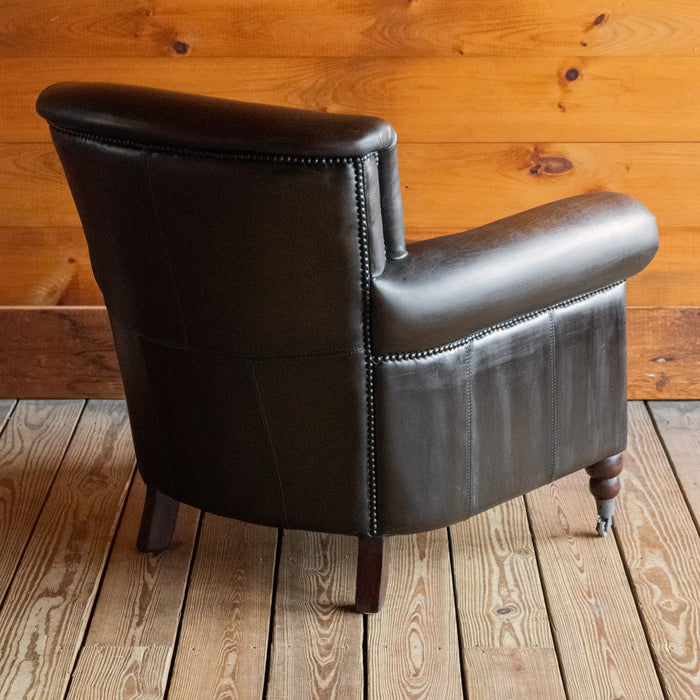 Angled back view of a rustic dark brown leather chair with rolled arms on a wooden floor against a wooden wall 