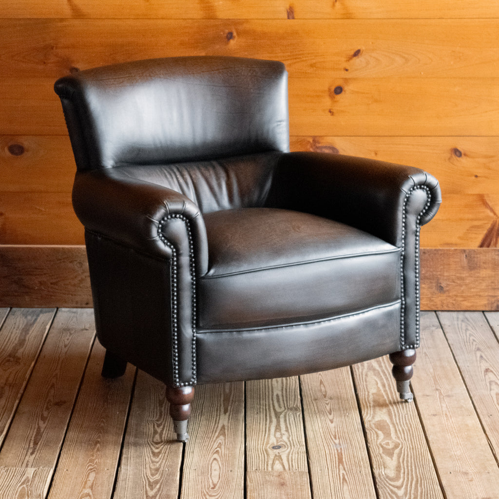 Angled front view of a rustic dark brown leather chair with rolled arms on a wooden floor against a wooden wall 