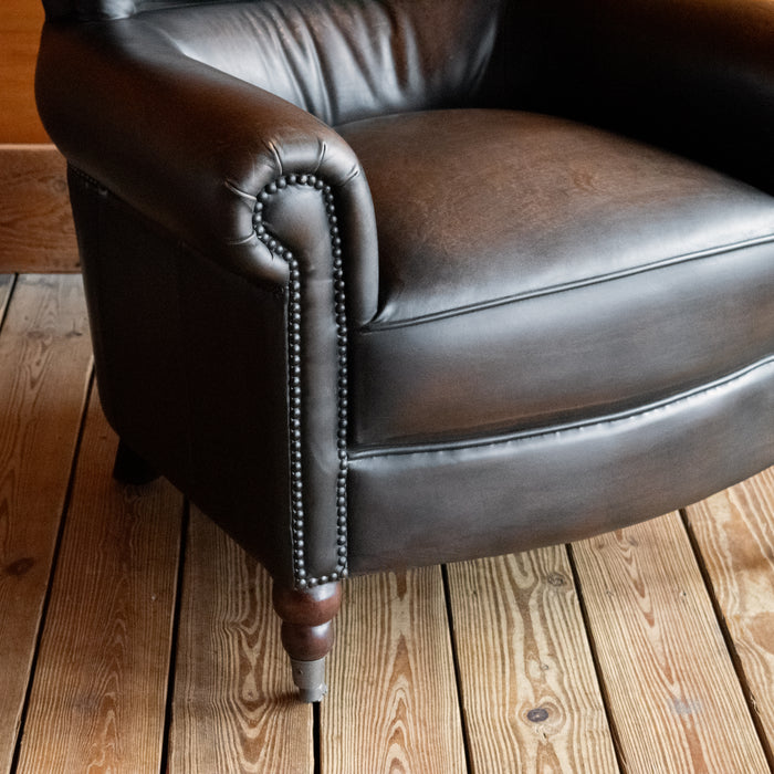Close-up of the arm of a rustic dark brown leather chair with rolled arms on a wooden floor