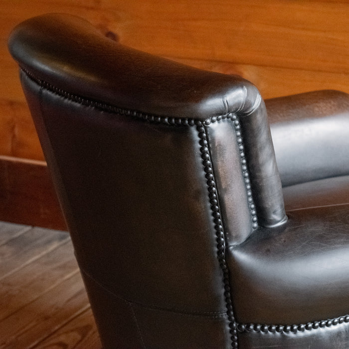 Close-up of the back corner of a rustic dark brown leather chair with rolled arms on a wooden floor against a wooden wall 