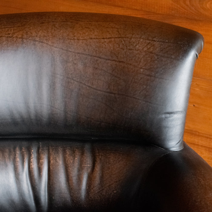 Close-up of the seatback of a rustic dark brown leather chair with rolled arms against a wooden wall 