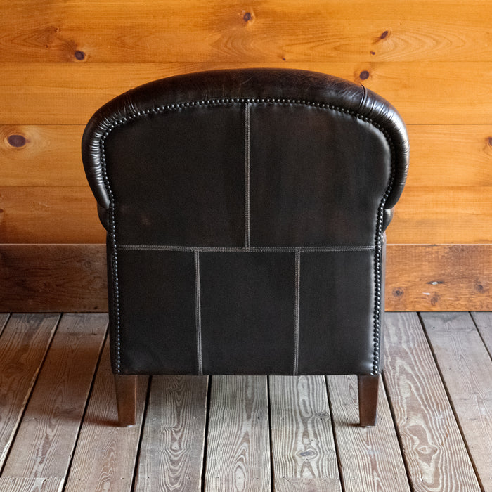Back view of a tight back rustic arm chair with nailhead trim against a wooden wall 