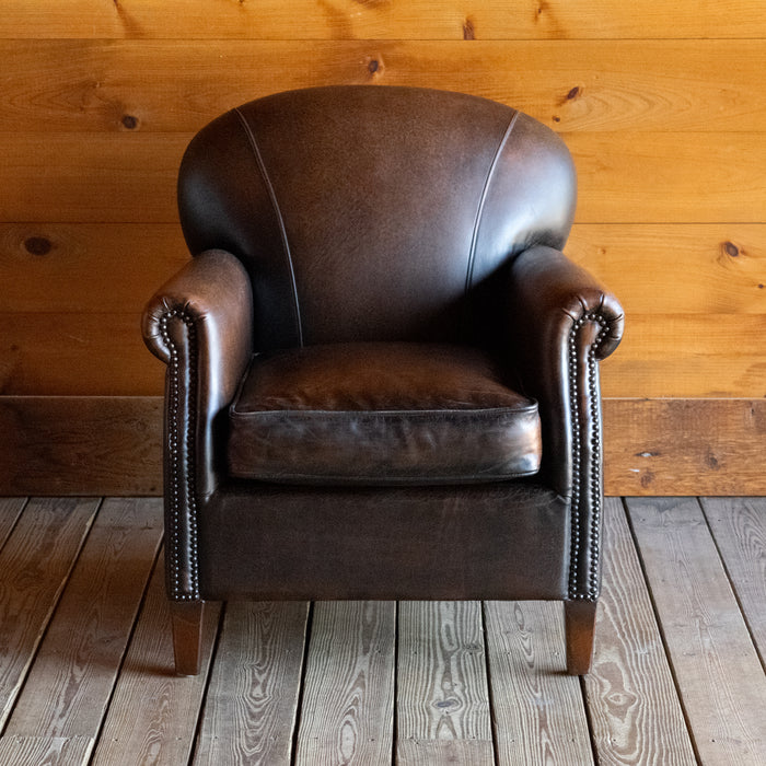Front view of a tight back rustic arm chair with nailhead trim against a wooden wall 