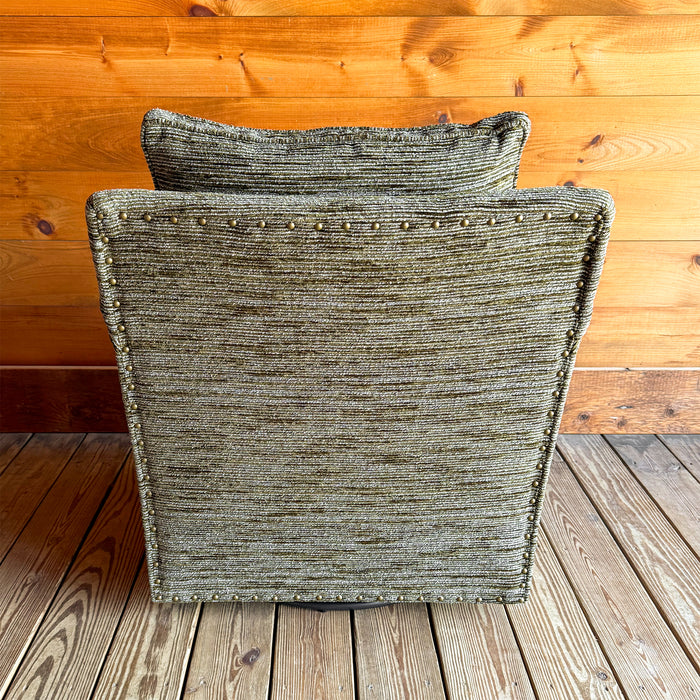 Back front view of a rustic track arm swivel chair upholstered in a mossy, mottled fabric against a wooden wall