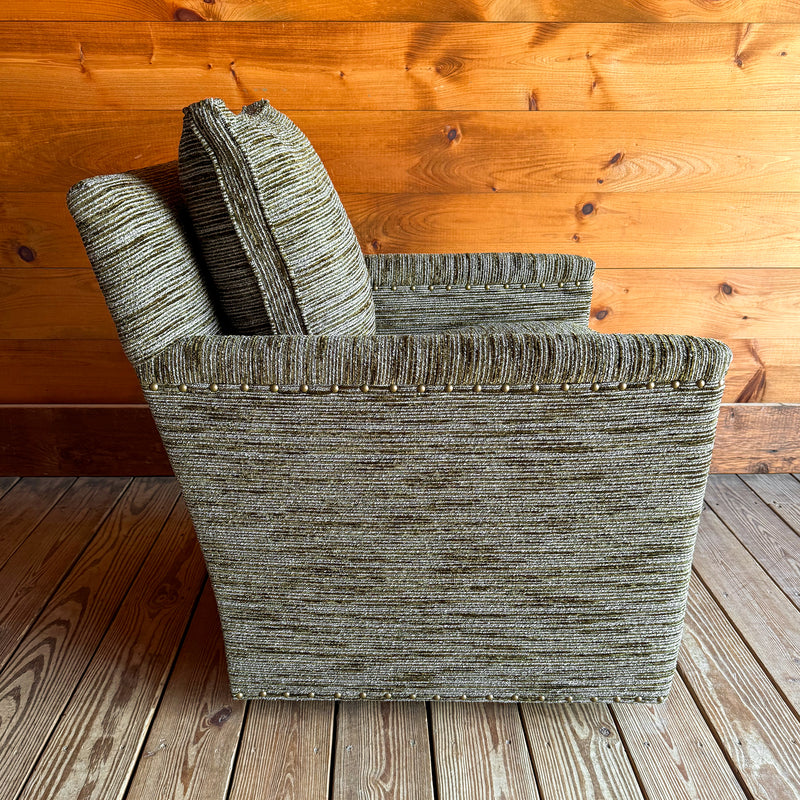 Profile view of a rustic track arm swivel chair upholstered in a mossy, mottled fabric against a wooden wall