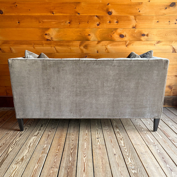 Back view of a brown-gray velvet tufted shelter arm sofa on a wooden floor against a wooden wall