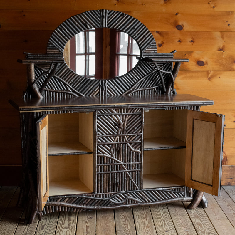Angled front view of an Adirondack style twig mosaic sideboard with open doors showing hidden storage, a gallery, mirror, and display shelves on a wide plank wooden floor against a rustic wooden wall