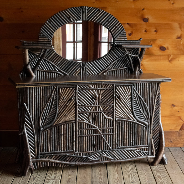 Angled front view of an Adirondack style twig mosaic sideboard with hidden storage, a gallery, mirror, and display shelves on a wide plank wooden floor against a rustic wooden wall