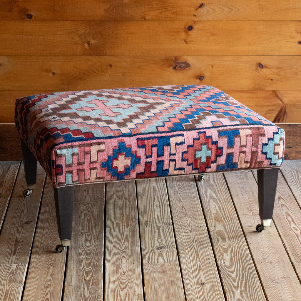 Angled front view of a vintage rug covered ottoman in shades of blue, rust, cream, and brown with tapered wooden legs on casters, on a wide-plank wooden floor against a rustic wooden wall