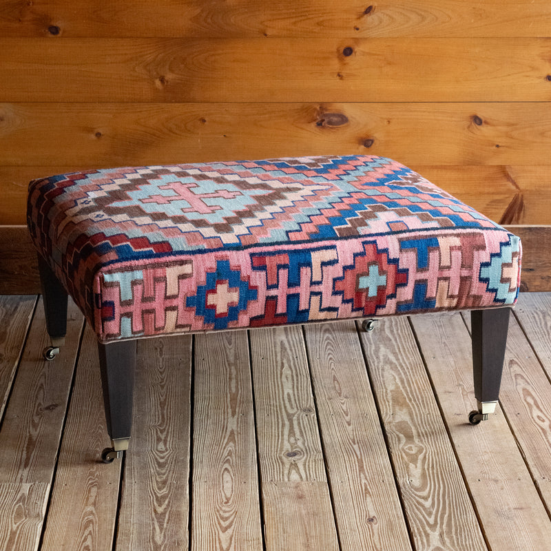 Angled front view of a vintage rug covered ottoman in shades of blue, rust, cream, and brown with tapered wooden legs on casters, on a wide-plank wooden floor against a rustic wooden wall