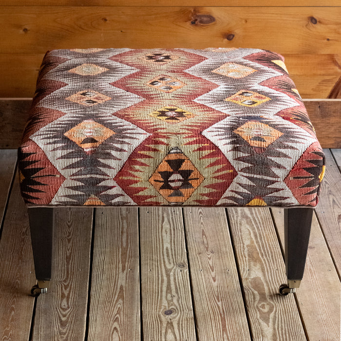 Front view of a vintage rug covered ottoman in shades of rust, green, cream, and brown with tapered wooden legs on casters, on a wide-plank wooden floor against a rustic wooden wall