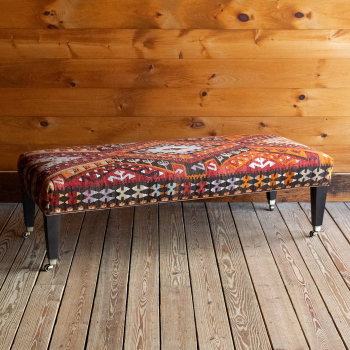 Angled front view of a vintage rug covered ottoman in shades of red, orange, cream, and brown with tapered wooden legs on casters, on a wide-plank wooden floor against a rustic wooden wall