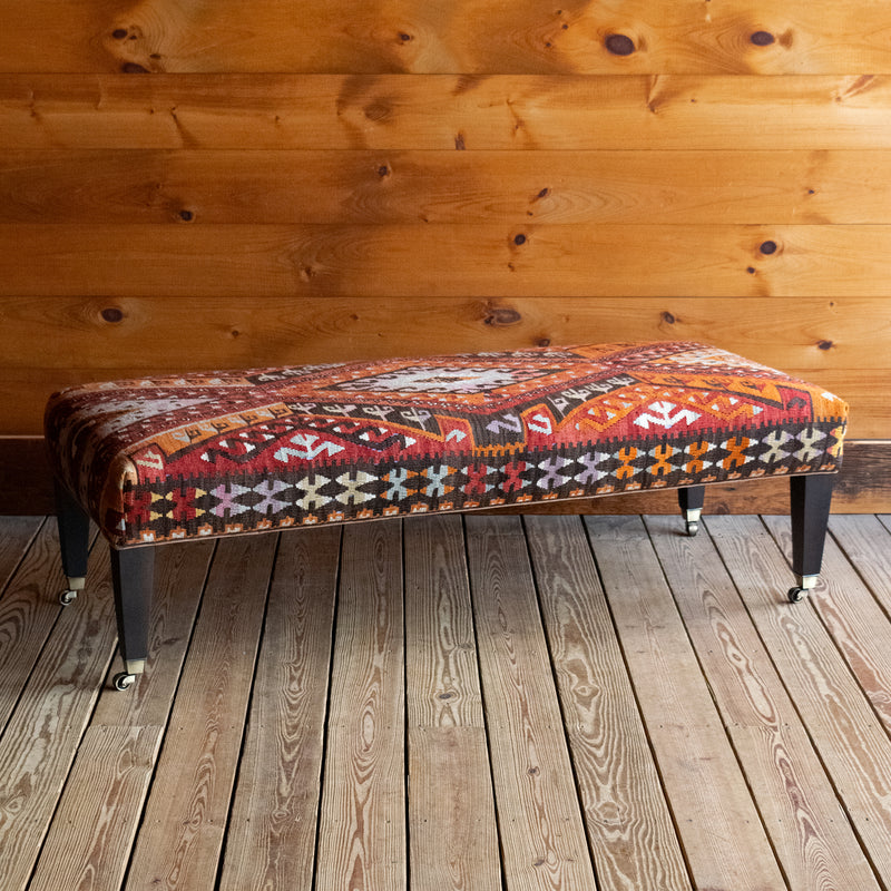 Angled front view of a vintage rug covered ottoman in shades of red, orange, cream, and brown with tapered wooden legs on casters, on a wide-plank wooden floor against a rustic wooden wall