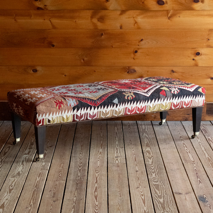Angled front view of a vintage rug covered ottoman in shades of red, blue, cream, and brown with tapered wooden legs on casters, on a wide-plank wooden floor against a rustic wooden wall
