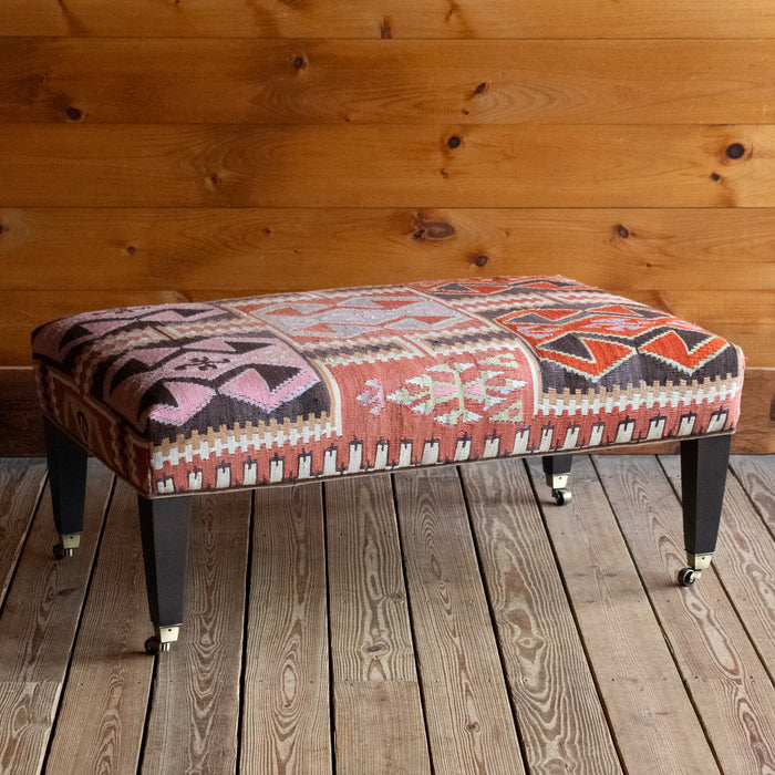 Angled front view of a vintage rug covered ottoman in shades of rust, green, cream, and brown with tapered wooden legs on casters, on a wide-plank wooden floor against a rustic wooden wall
