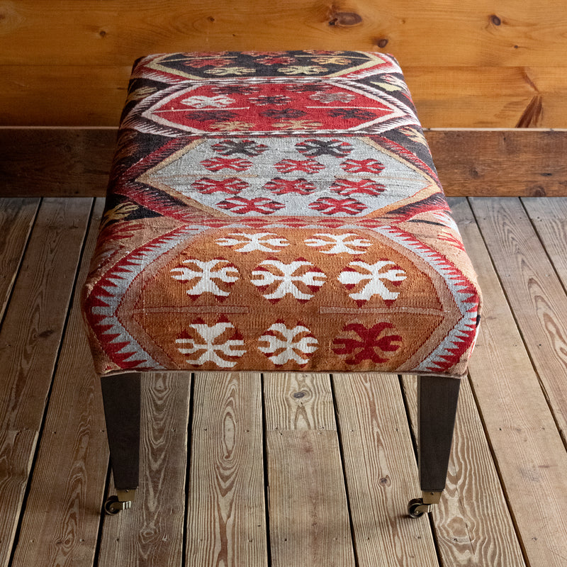 Front view of a vintage rug covered ottoman in shades of red, blue, cream, and brown with tapered wooden legs on casters, on a wide-plank wooden floor against a rustic wooden wall
