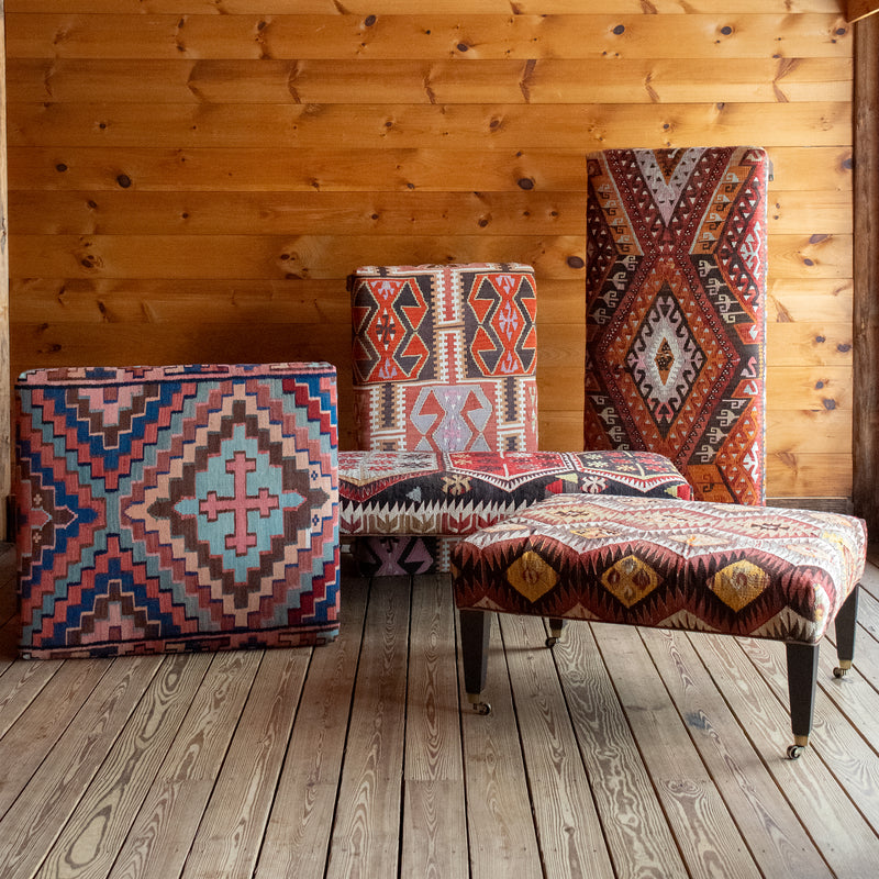 Five vintage rug covered ottomans on a wide-plank wooden floor against a rustic wooden wall