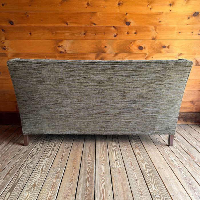Back view of a 75" wingback apartment sofa upholstered in a mossy, mottled fabric against a wooden wall