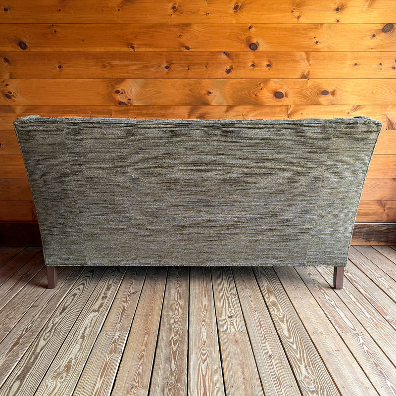 Back view of a 75" wingback apartment sofa upholstered in a mossy, mottled fabric against a wooden wall