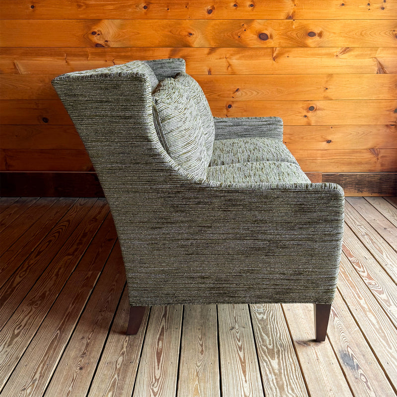 Profile view of a 75" wingback apartment sofa upholstered in a mossy, mottled fabric against a wooden wall
